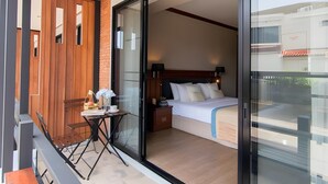 Una habitación de hotel moderna con un amplio ventanal corredero que da a un balcón, una cama bien hecha con cabecera de madera, y una mesita con una silla en el balcón.
