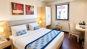 A hotel room with a large bed, bedside tables, a desk, and a chair. There are two paintings on the wall, a window with a view, and a door. - The Theme Jaipur (Jaipur)