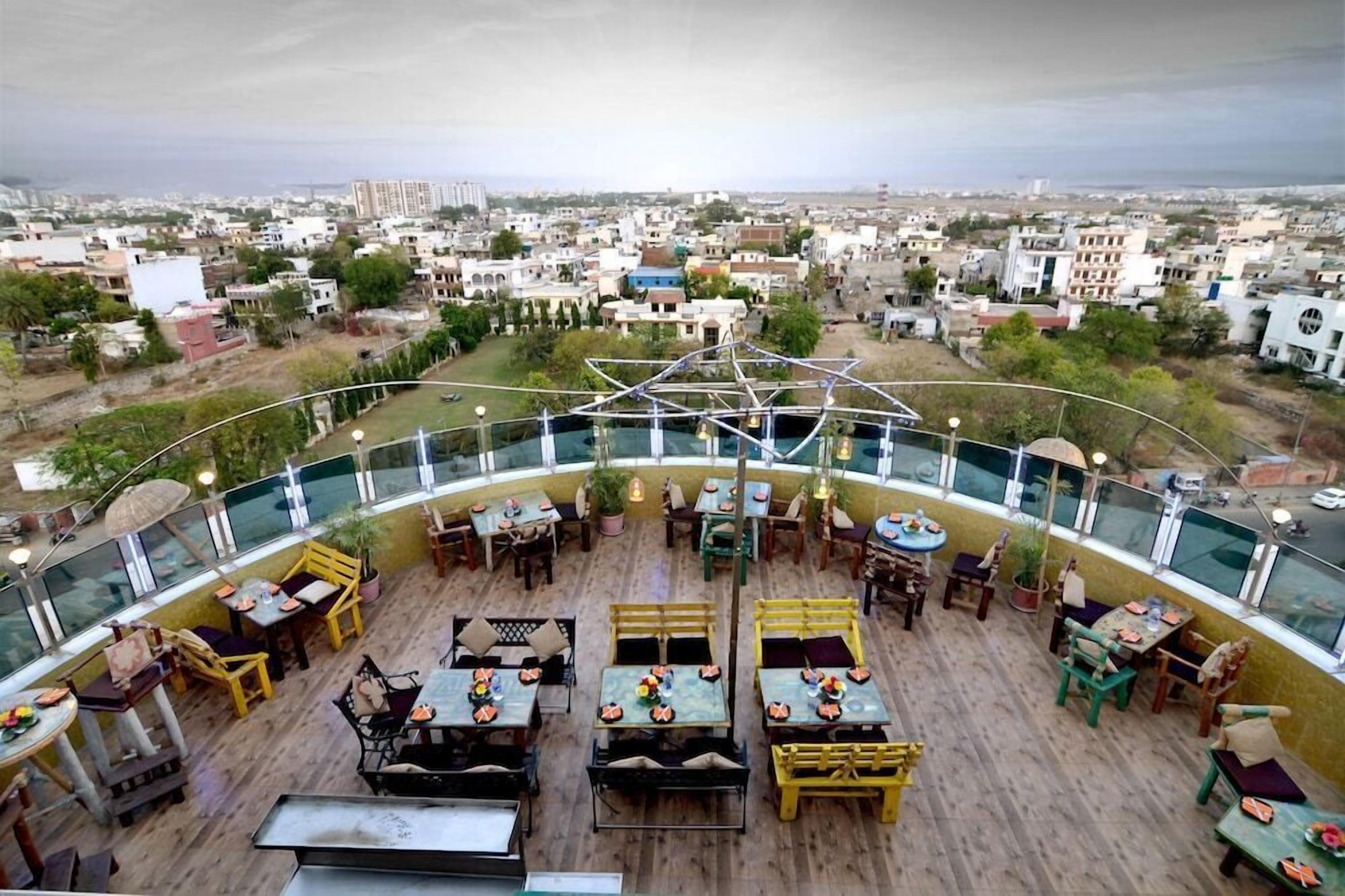 An outdoor dining area with tables and chairs, surrounded by a glass railing, overlooking a cityscape.