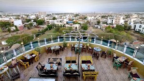 An outdoor dining area with tables and chairs, surrounded by a glass railing, overlooking a cityscape. - The Theme Jaipur (Jaipur)
