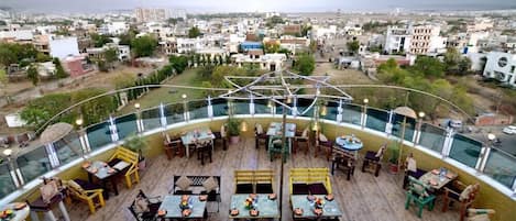 An outdoor dining area with tables and chairs, surrounded by a glass railing, overlooking a cityscape.