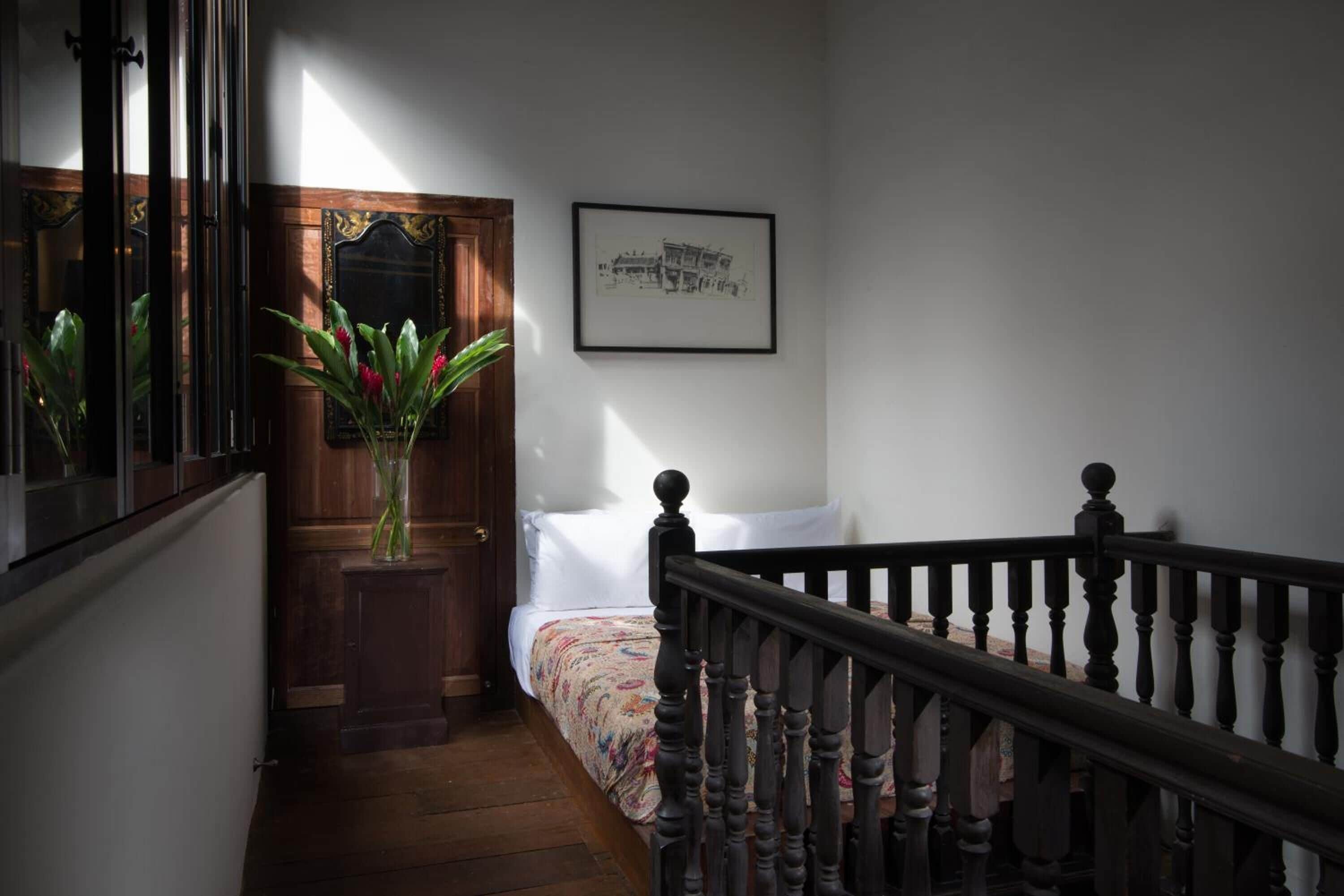 A narrow hallway with a wooden door, a bed with floral bedding, a framed picture on the wall, and a staircase with a wooden railing.