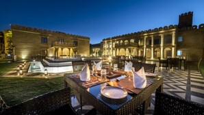 Un área de comedor al aire libre con sillas y mesas de mimbre, una fuente y un edificio con ventanas arqueadas al fondo.