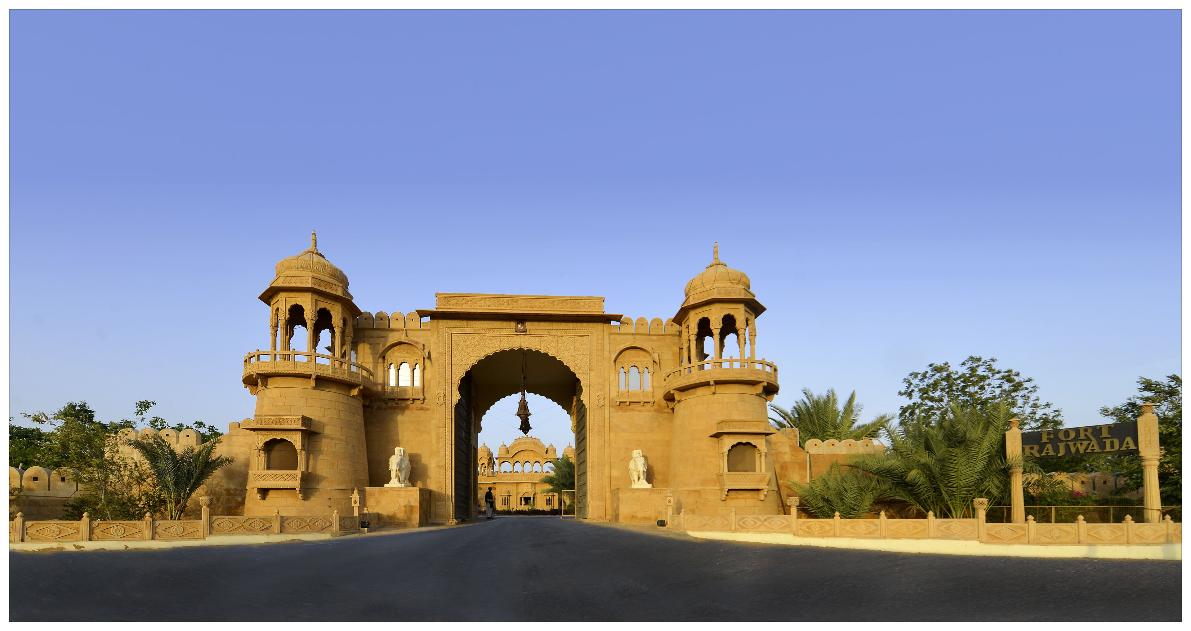 A large, ornate archway with two towers and a central dome, leading to a building with a sign reading "Fort Rajwada".