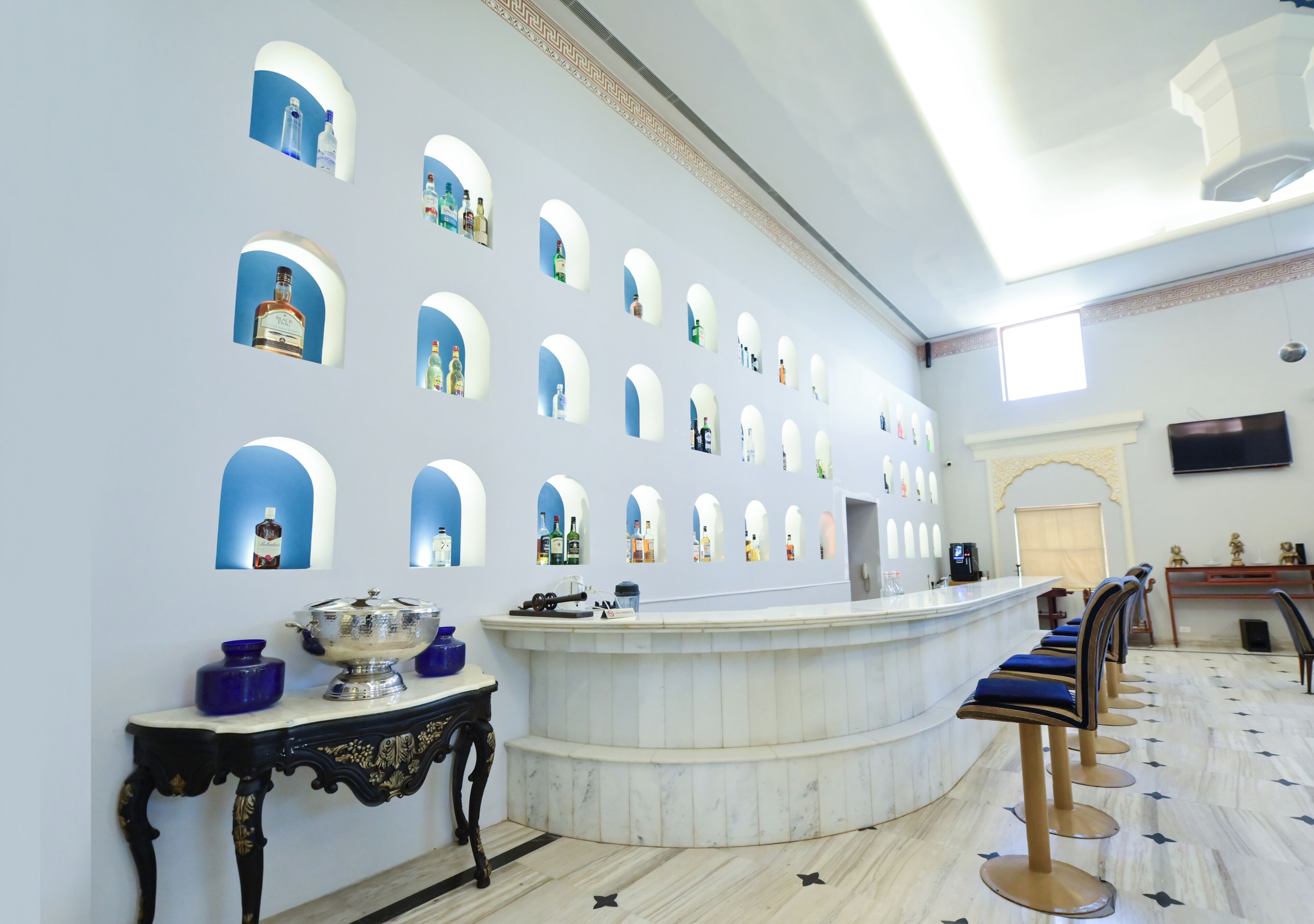A bar area with a marble counter, blue bar stools, and a wall with multiple small windows displaying bottles.