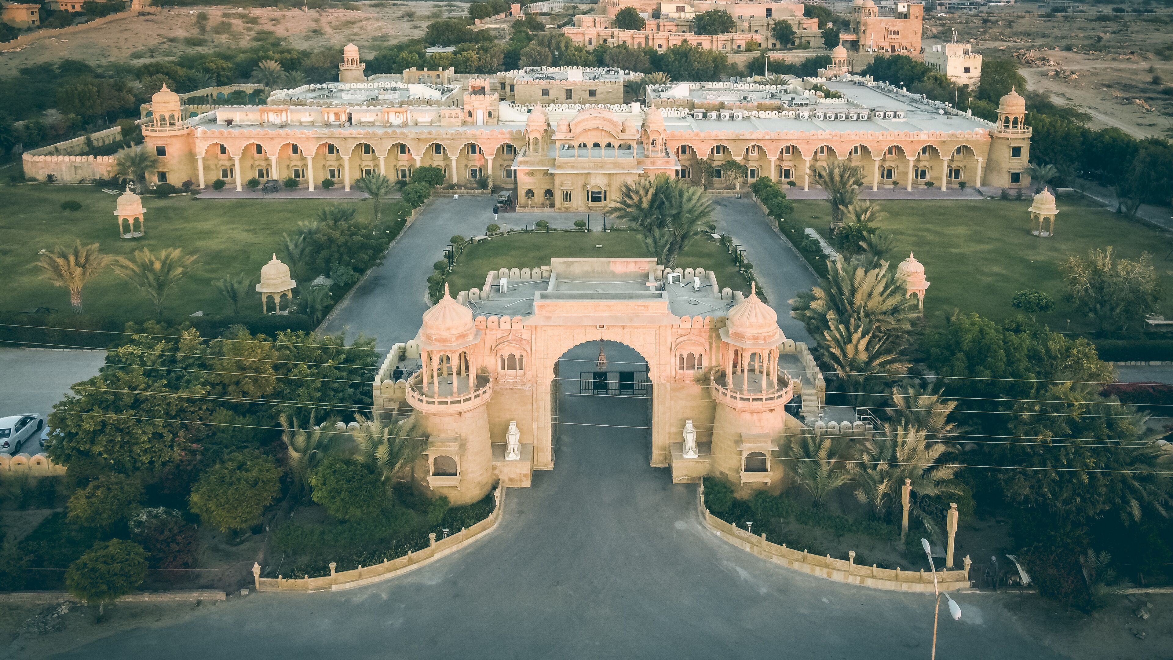 Vue aérienne d’un grand bâtiment historique doté d’une arche centrale, entouré d’une végétation luxuriante et de palmiers.
