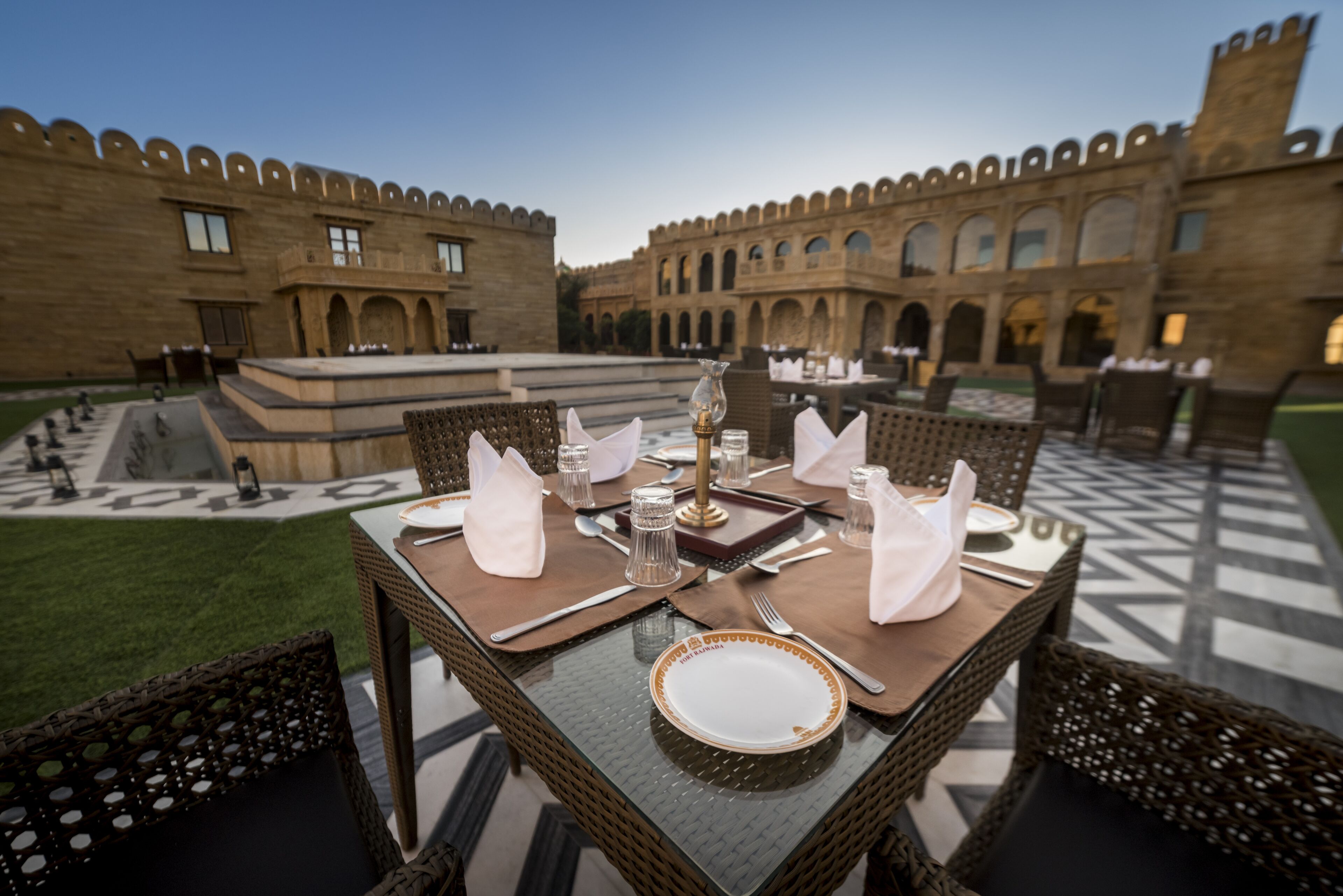 A dining table set with white plates, silverware, and napkins, surrounded by wicker chairs, with a courtyard and historic building in the background.