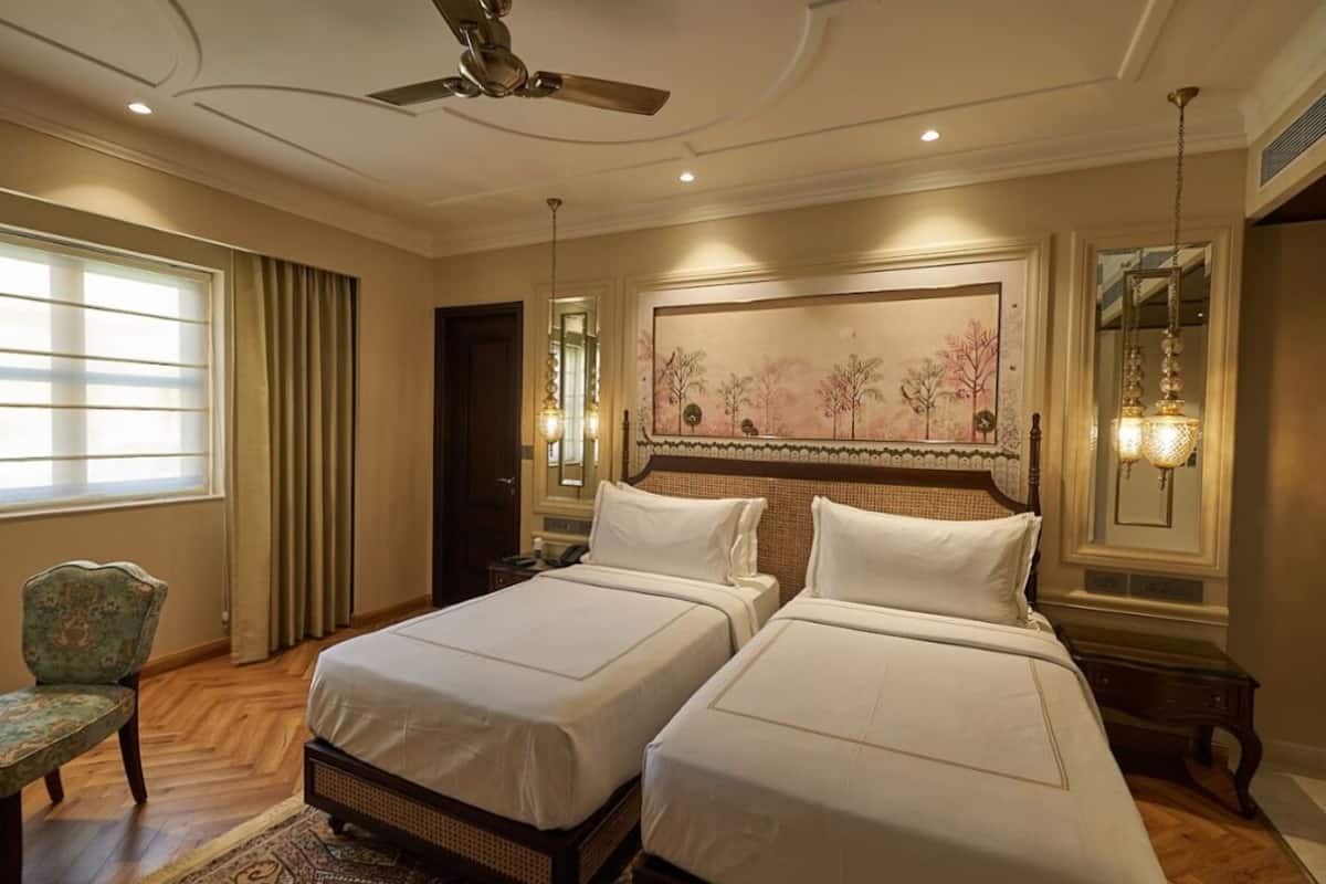 A hotel room with two beds, a ceiling fan, a large painting, and a chair.