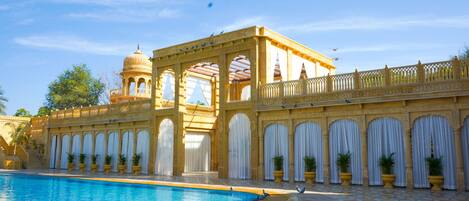 Un área de piscina con suelo de baldosas, un edificio con arcos y una cúpula, y plantas en macetas a lo largo del sendero.