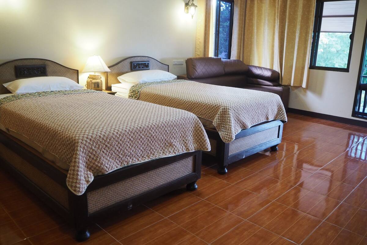 A hotel room with two beds, a nightstand, a lamp, a window with curtains, and a view of greenery outside.