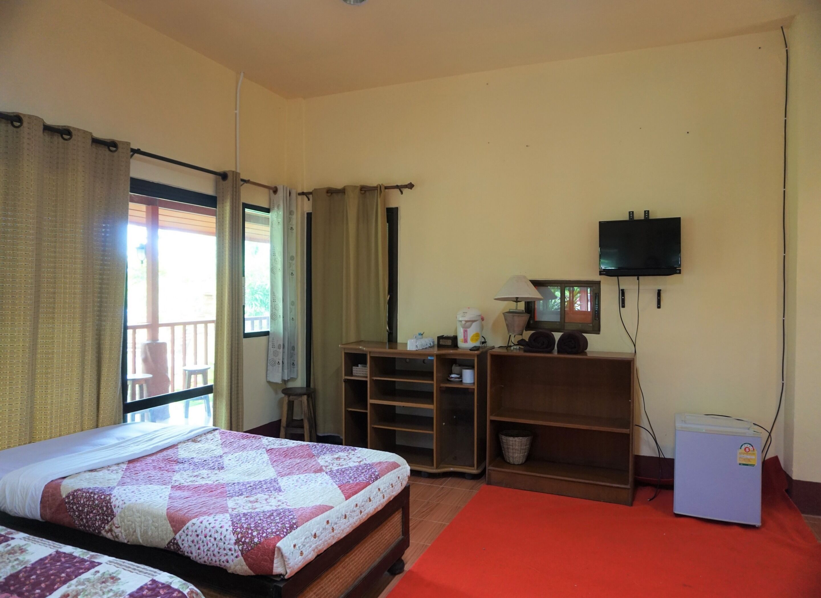 A bedroom with a bed, a television, a small refrigerator, and a desk with a lamp.