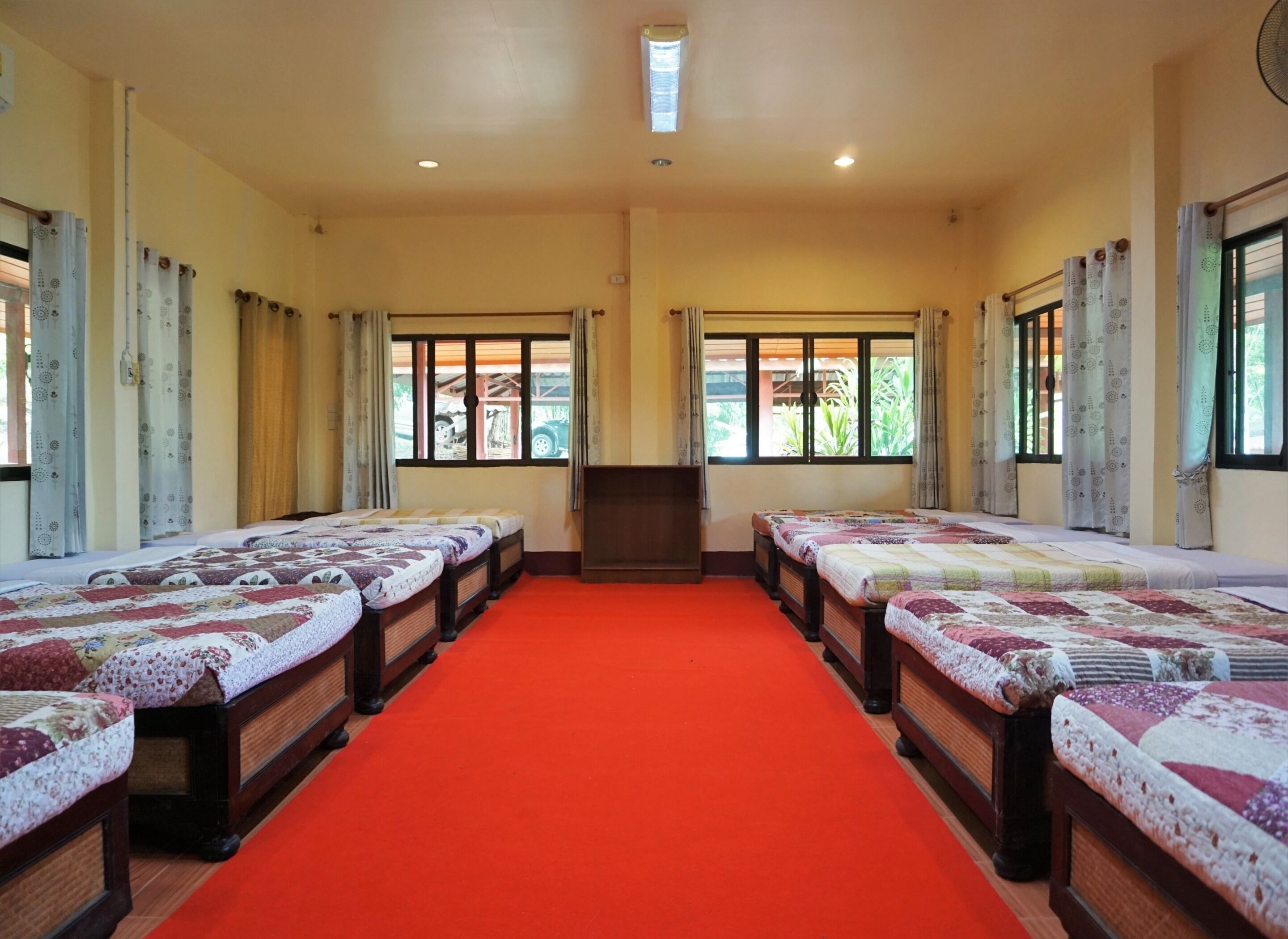 A hotel room with four beds aligned along a red carpet, large windows with curtains, and a ceiling fan.