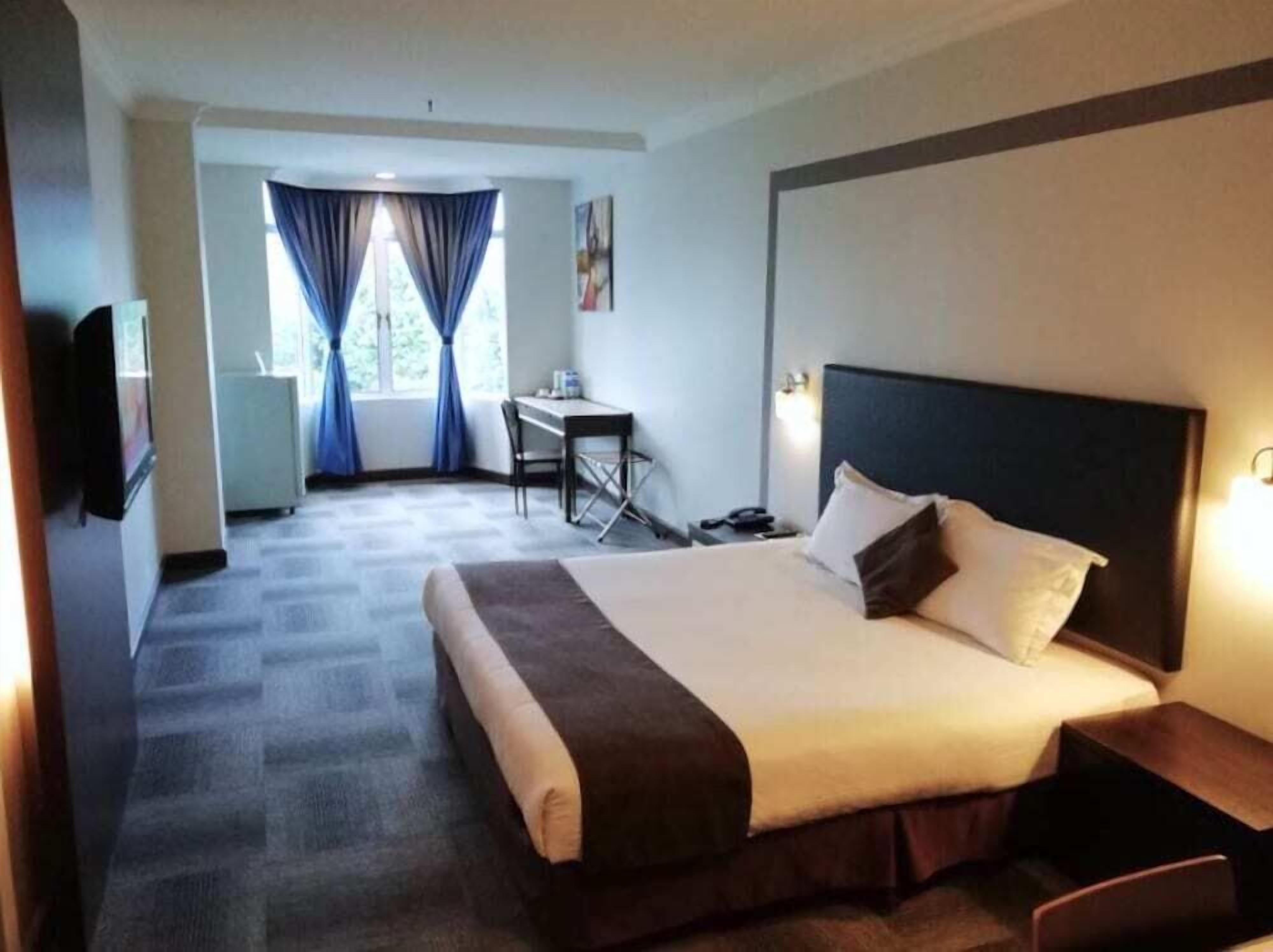 A hotel room with a large bed, a desk, and a chair. There is a window with curtains, a television, and a wall-mounted light.