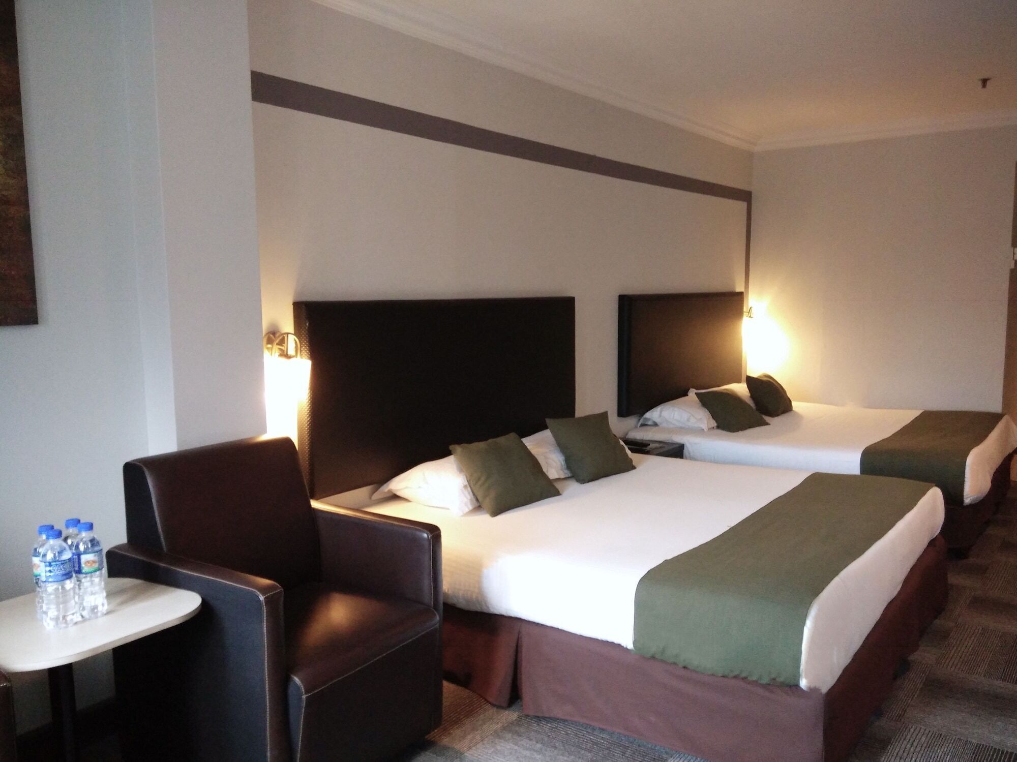 A hotel room with two beds, a small table with water bottles, and a leather chair.