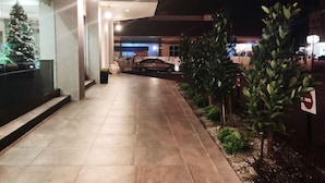 Vista nocturna del exterior de un edificio con un camino pavimentado, un coche aparcado y un árbol de Navidad en la ventana.