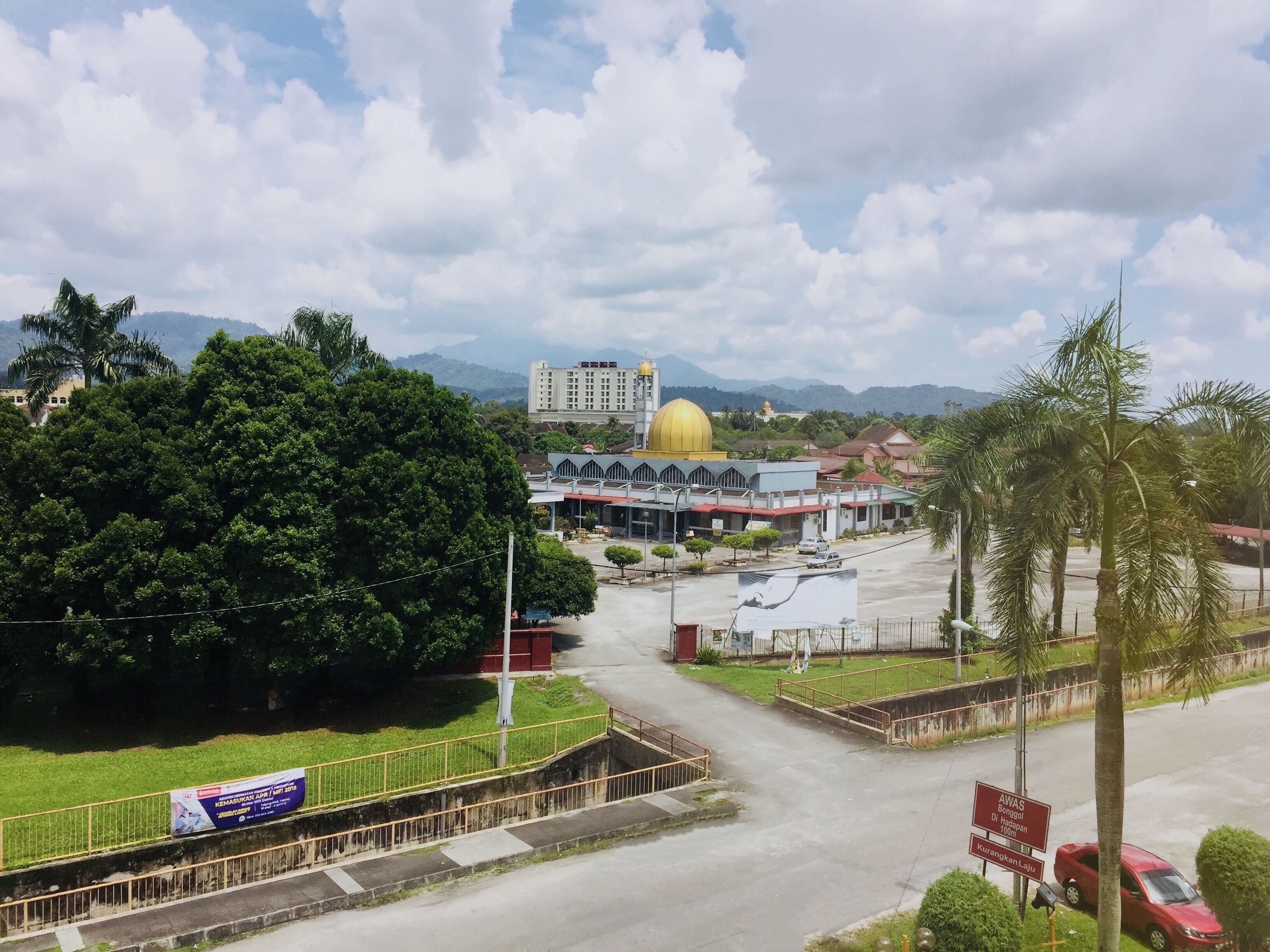 A view of a building with a large golden dome, surrounded by greenery and a road.