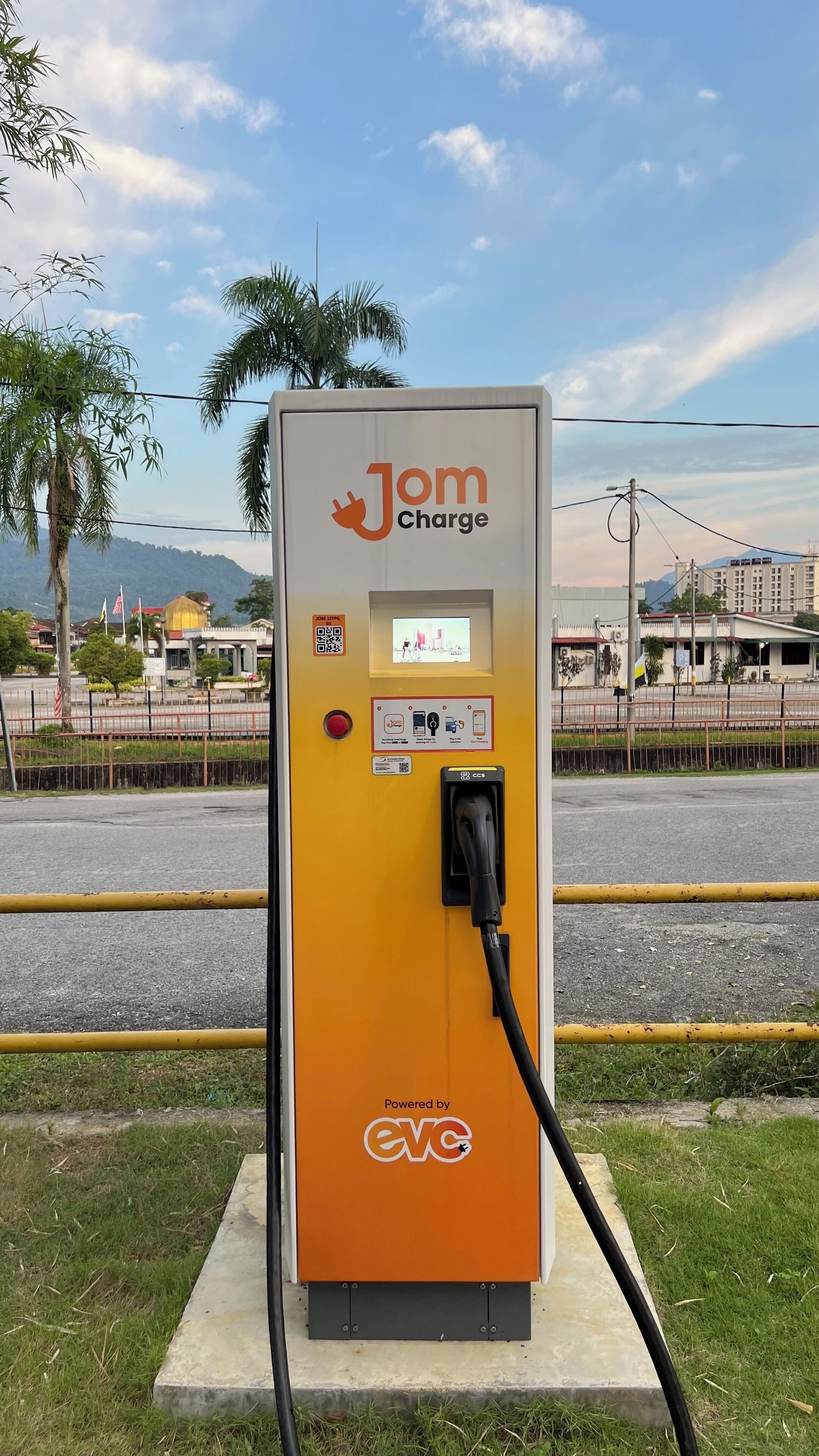 A Jom Charge electric vehicle charging station with a digital display, a QR code, and a CCS connector.