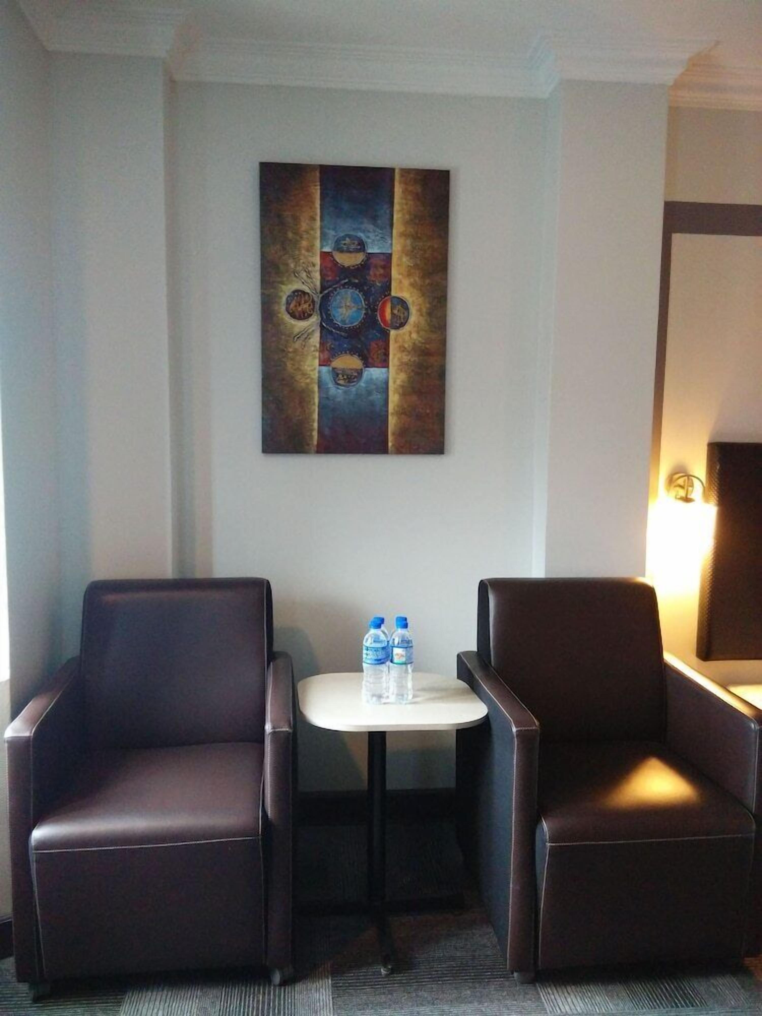 A hotel room with two leather armchairs, a small table with two water bottles, and a large abstract painting on the wall.