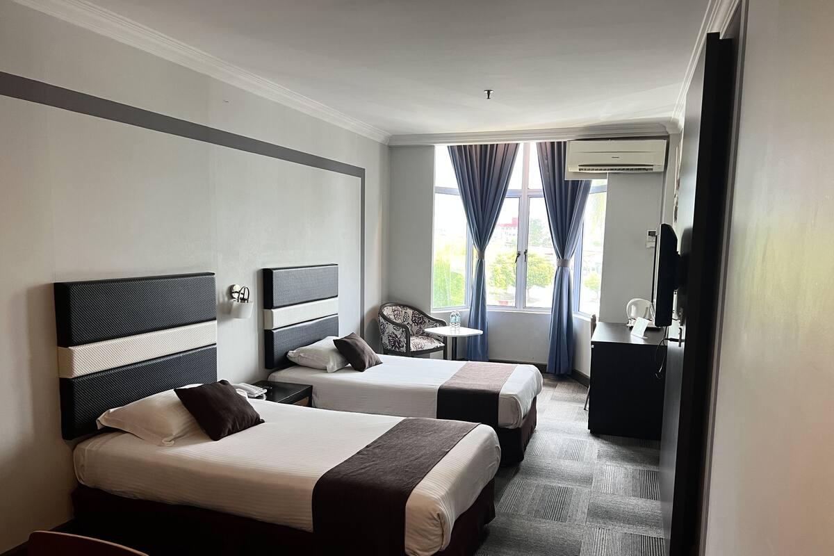 A hotel room with two beds, a desk, a chair, a small table, and an air conditioning unit.