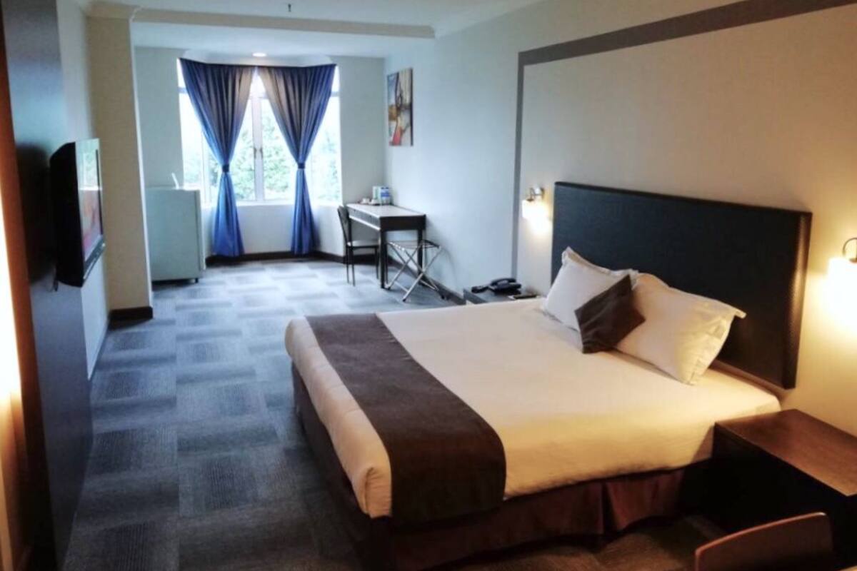 A hotel room with a large bed, a desk, and a chair. There is a window with curtains, a flat-screen TV, and a wall-mounted light.