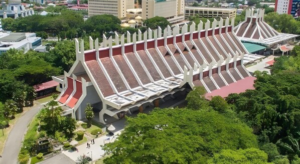 A large, modern amphitheater with tiered seating and a central stage, surrounded by lush greenery and urban buildings.