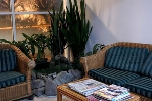 Una habitación con muebles de mimbre, una mesa de centro con revistas y plantas verdes grandes.