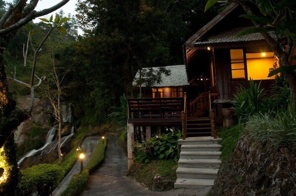 A wooden cabin with a staircase leading to a porch, surrounded by lush greenery and a waterfall. - Kangsadarn Resort & Waterfall (Chiang Mai)