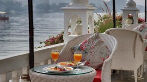 A dining table with plates of food and glasses of orange juice, overlooking a body of water with boats and buildings in the background. - Lake Pichola (Udaipur)