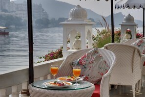 A dining table with plates of food and glasses of orange juice, overlooking a body of water with boats and buildings in the background. - Lake Pichola (Udaipur)