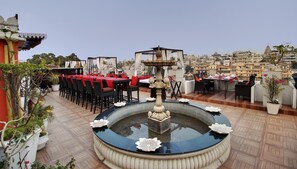 A rooftop dining area with tables and chairs, a decorative fountain, and a view of the cityscape. - Lake Pichola (Udaipur)