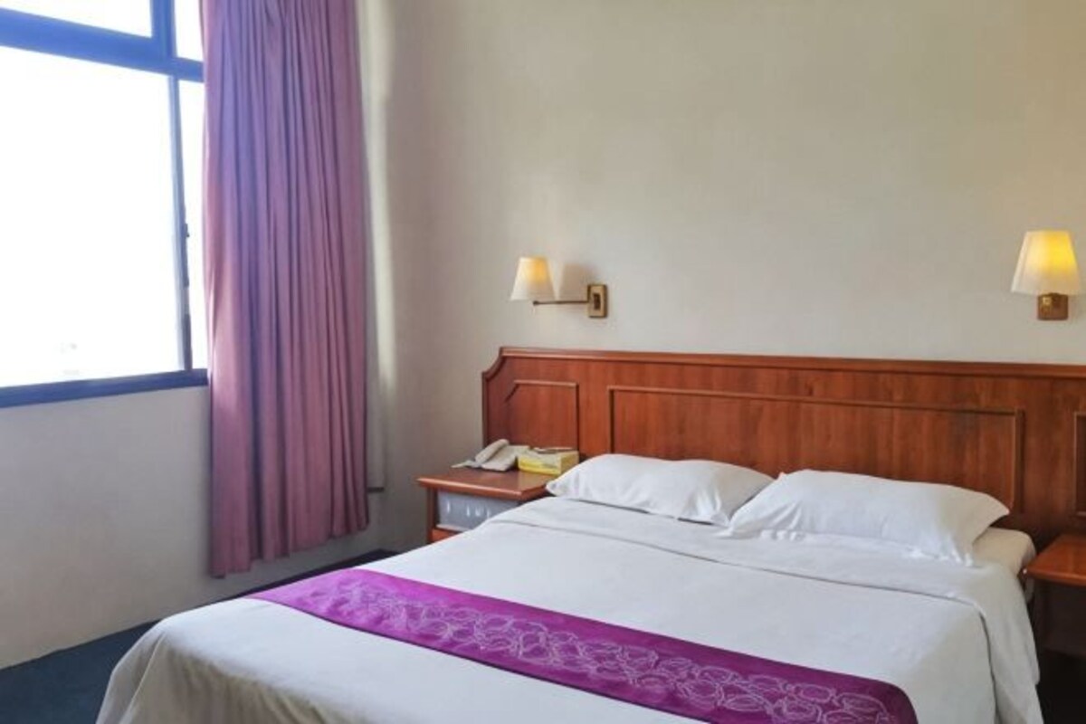 A hotel room with a wooden headboard, a bed with white linens, a nightstand with a lamp, and a window with curtains.