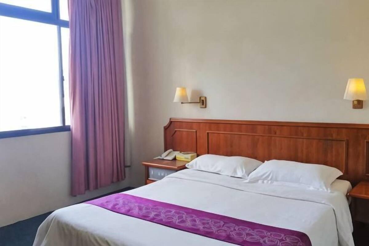 Camera d'albergo con testiera in legno, letto con lenzuola bianche, comodino con lampada e finestra con tende.