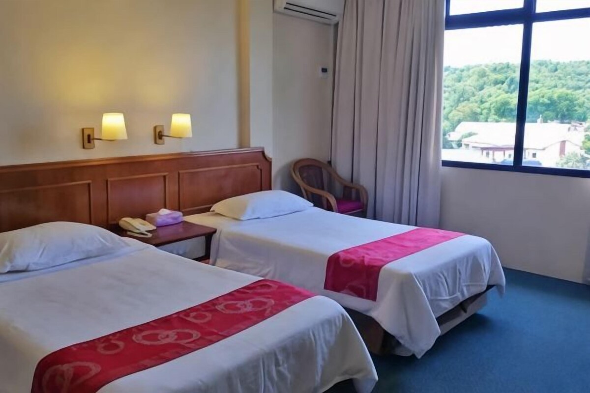 Hotelkamer met twee bedden, een houten hoofdbord, een nachtkastje met telefoon en uitzicht op groen.
