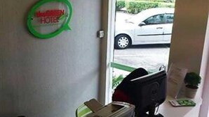 Un comptoir d'accueil avec une enseigne verte indiquant qu'il s'agit d'un hôtel, un ordinateur et une fenêtre donnant sur une voiture garée à l'extérieur.