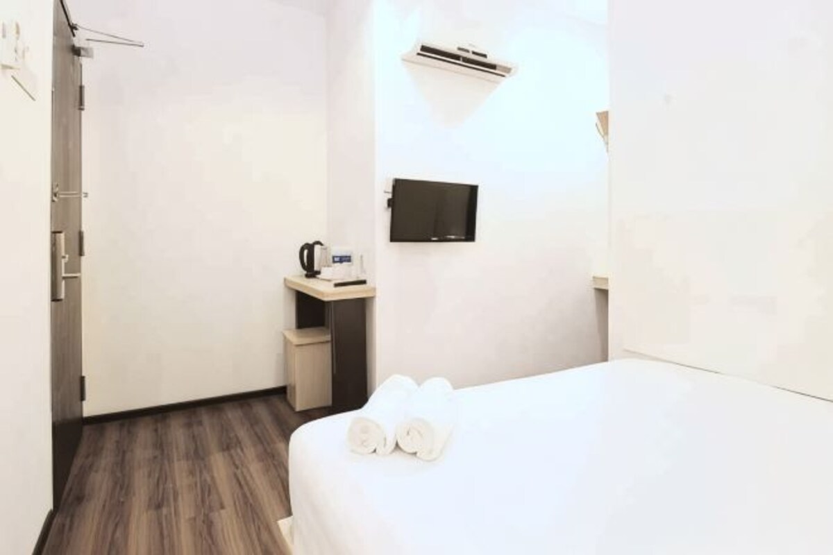 A hotel room with a bed, a small desk with a kettle, a TV, and a wall-mounted air conditioner.