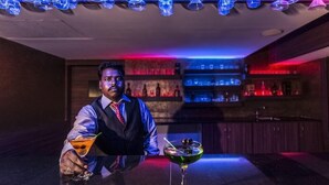 A bar area with a bartender, a bar counter, and shelves with bottles in the background. - Clinton Park Inn (Nagapattinam)