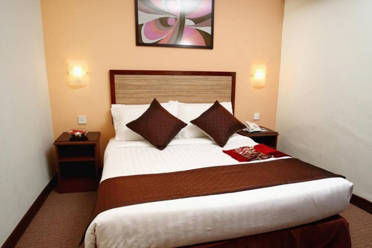 A neatly made bed with a wooden headboard, two pillows, a red throw blanket, and a nightstand with a phone and a small table with a bowl of fruit.