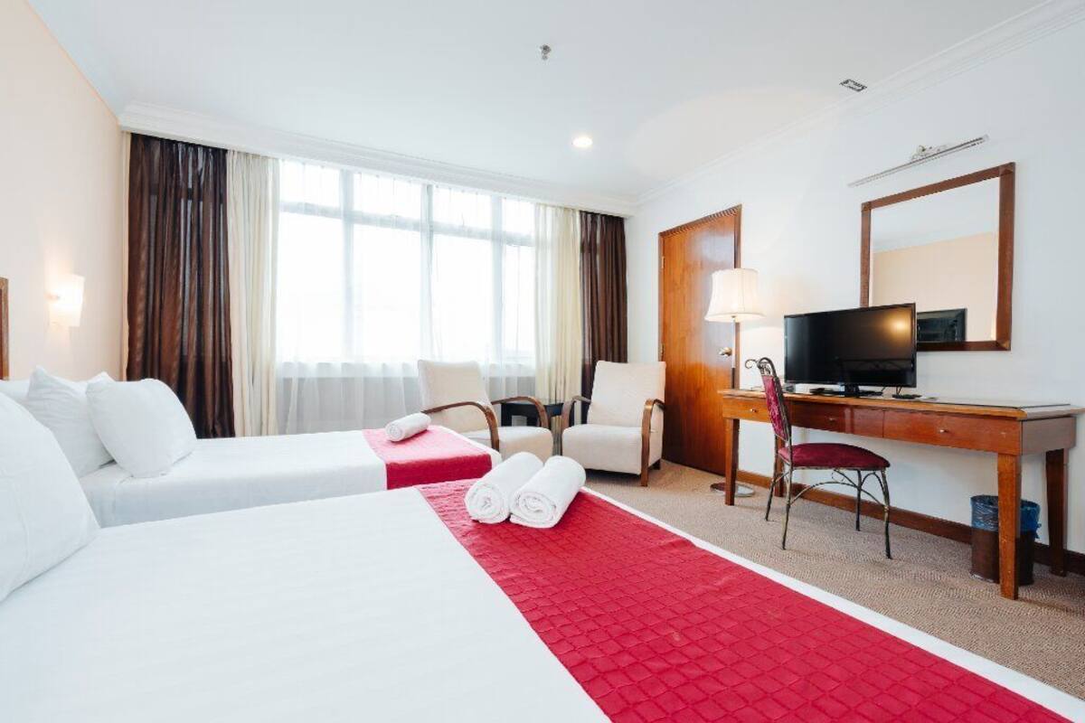 A hotel room with two beds, a desk with a TV, a chair, and a large window with curtains.
