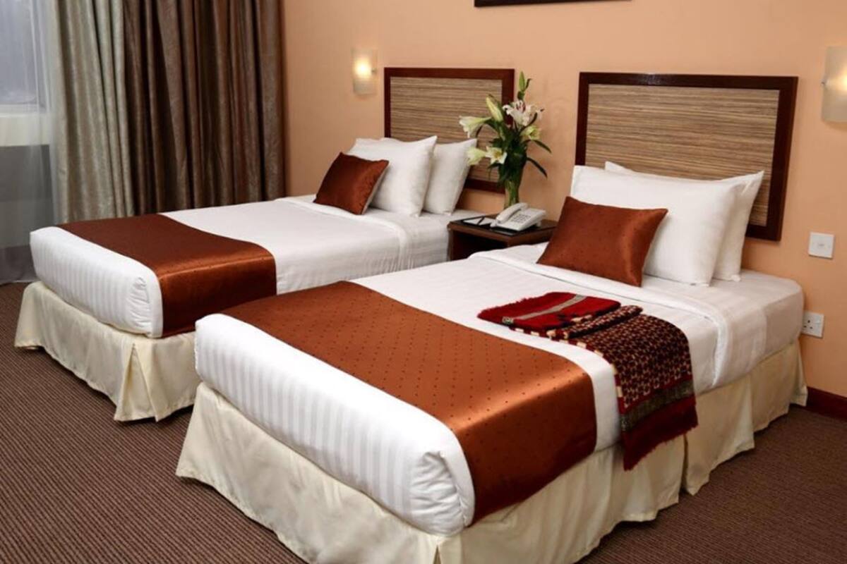 A hotel room with two beds, each with a brown headboard, white bedding, and a red throw blanket. There is a nightstand with a phone and a vase with flowers.