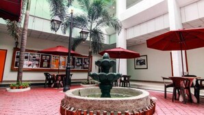 Un amplio patio interior con una fuente central, rodeado de palmeras y sombrillas rojas.
