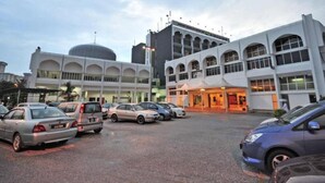 Un estacionamiento con varios coches, un edificio con cúpula y ventanas de arco, y otro con fachada plana.