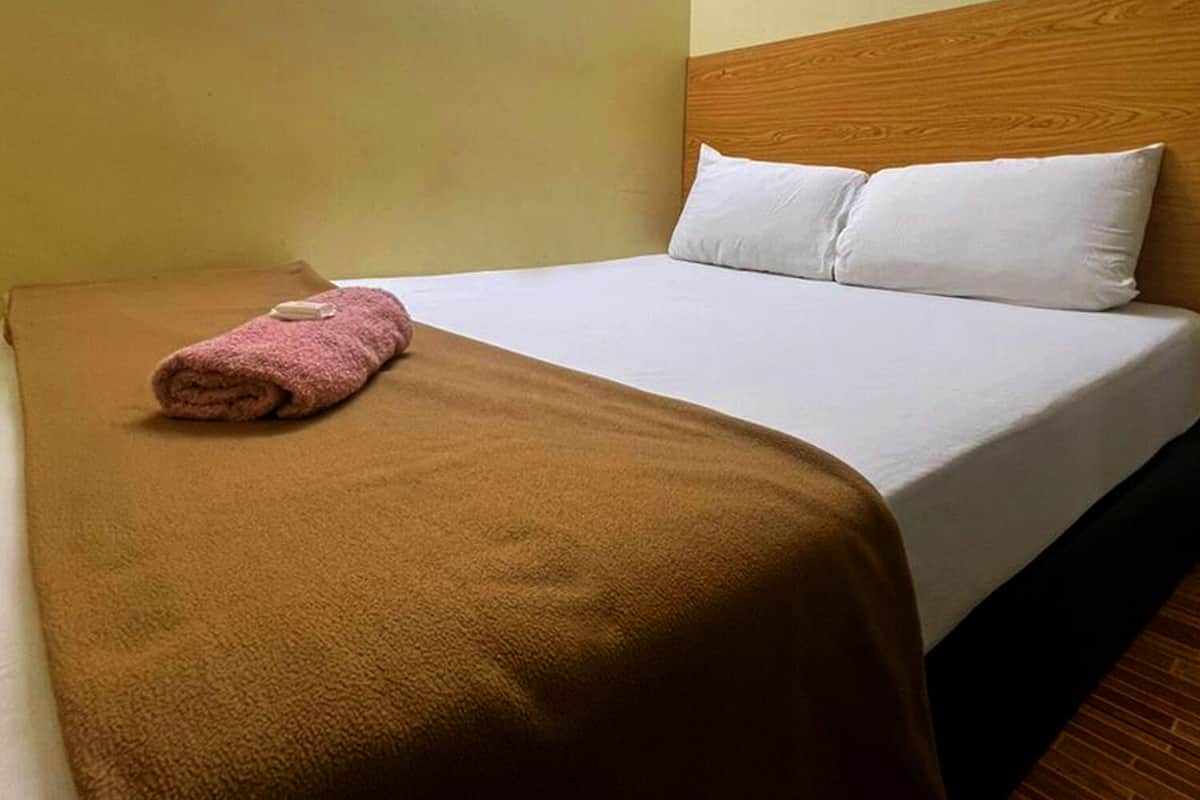 A single bed with a brown blanket and a pink towel, a wooden headboard, and a fluorescent light fixture above.