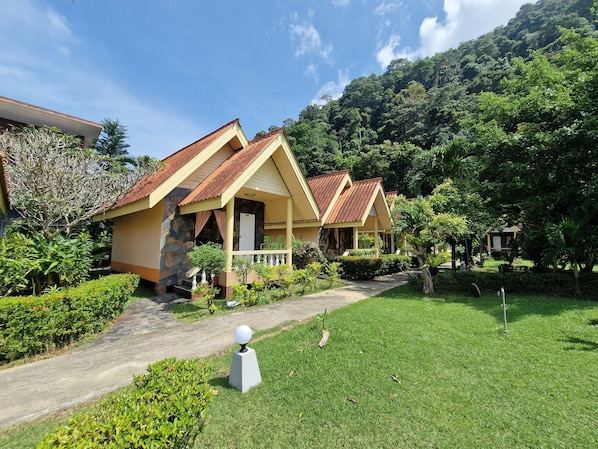 A row of yellow buildings with red roofs, surrounded by lush greenery and a mountain backdrop.