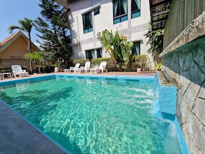 A swimming pool with clear water, surrounded by lounge chairs and potted plants, with a building in the background.