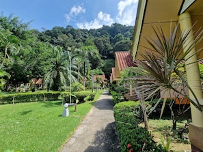 A pathway leading through a well-maintained garden with various plants and a building with a red roof in the background.
