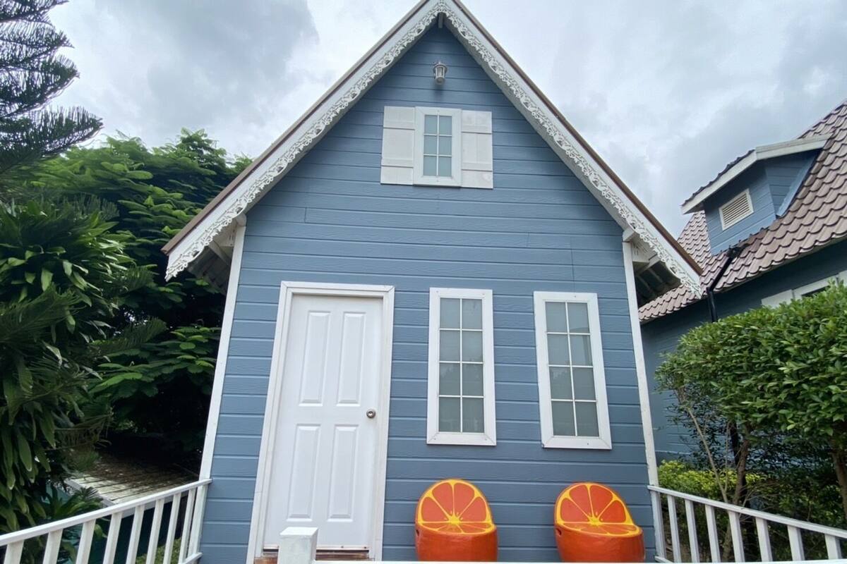 A blue house with a white door and two windows, a porch with a railing, and two orange planters.