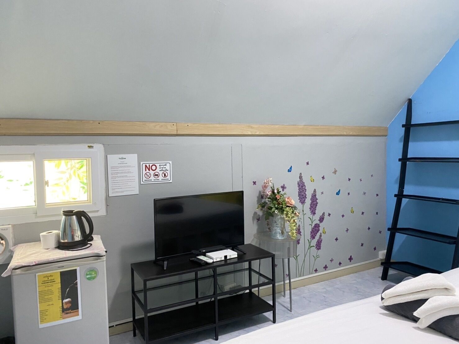 A room with a TV, a small table with a vase, a black shelf, and a window with blinds.