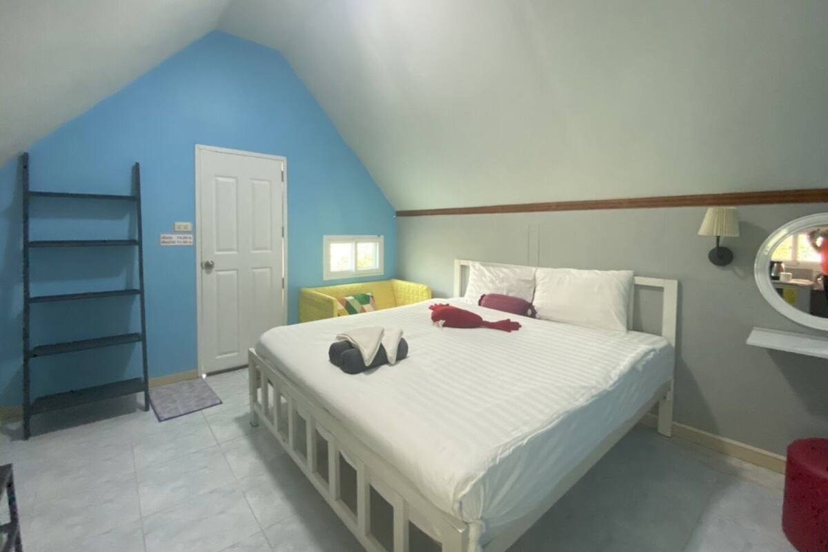 A bedroom with a sloped ceiling, a bed with white linens, a ladder, a small window, and a mirror on the wall.