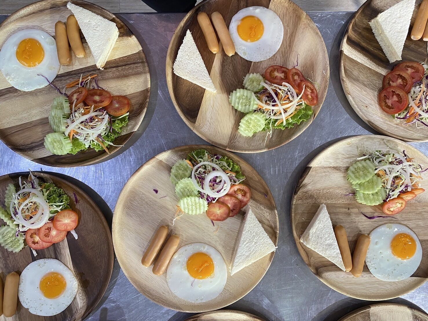 Una mesa con varios platos de madera, cada uno con un huevo frito, salchichas y una ensalada de tomates, pepinos y lechuga desmenuzada.