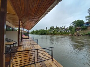 A wooden deck with metal railings overlooking a river, with buildings and greenery in the background. - Riverside Hill Resort & Restaurant (Sai Yok)